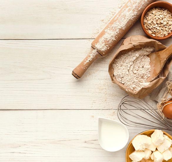 Cooking ingredients background. Border of flour, eggs, raisins, sugar, milk, butter and kitchen utensils on white rustic wood with copy space. Dough preparing and pastry concept, top view