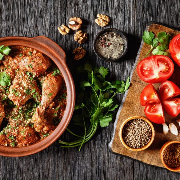 Chakhokhbili, stewed chicken, tomato with fresh herbs, walnuts and spices in a clay pot on a wooden table with ingredients on a cutting board, georgian dish, flat lay