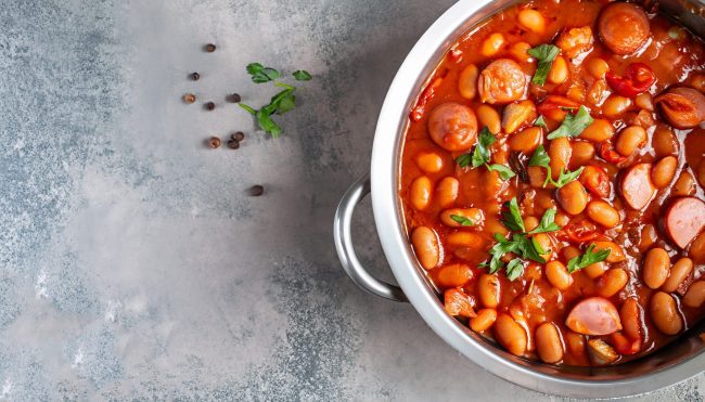 hearty beans stew with sausages, herbs and spices in tomato sauce in a metal casserole on a concrete table, fasolka po bretonsku, polish cuisine, view from above, flatllay, copy space