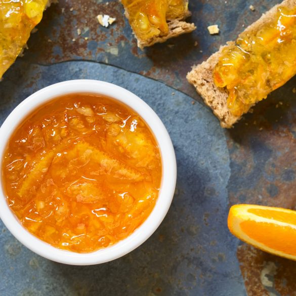 Orange jam in small bowl with bread and orange wedges on the side, photographed overhead on slate with natural light (Selective Focus, Focus on the jam in the bowl)