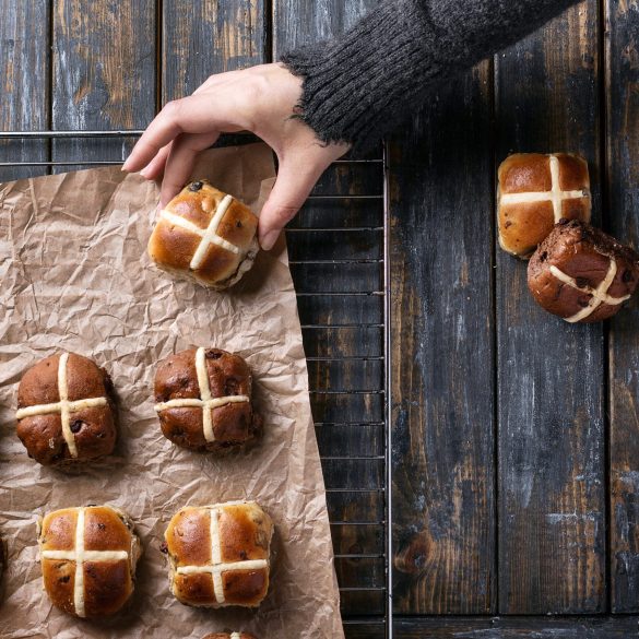 Traditional Easter treats cross buns with raisins and female hands on the grey wooden board. Top view