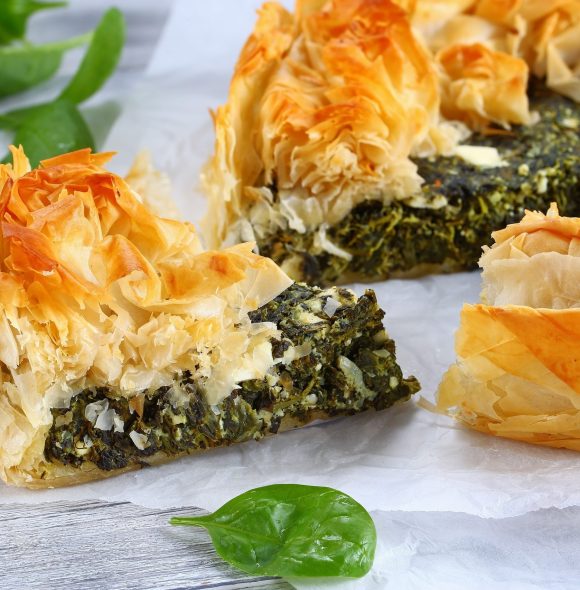 delicious golden crust hot greek spinach feta cheese pie or spanakopita cut in slices on white paper with spatula on table, authentic recipe, side view from above, close-up