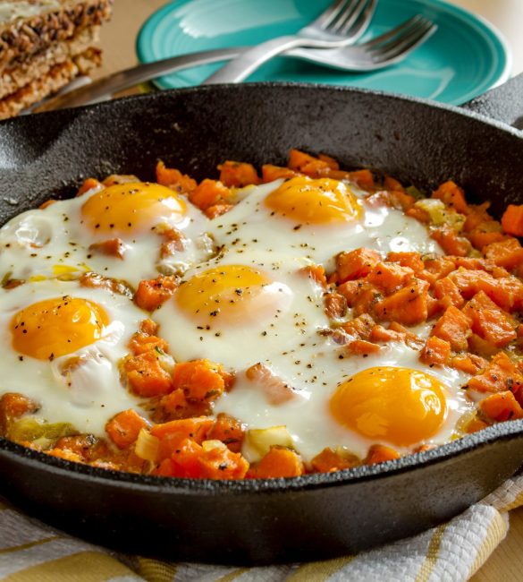Fried eggs and sweet potato hash in cast iron skillet sitting on yellow striped kitchen towel with whole grain toast and stacked plates