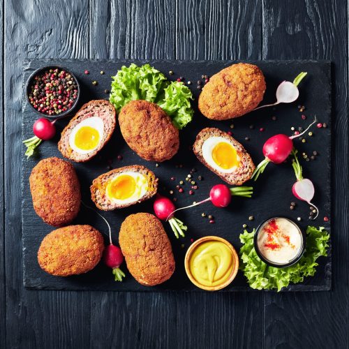 meatballs stuffed with soft boiled eggs, scotch eggs served with radish, mayonnaise and mustard on a black stone plate, view from above