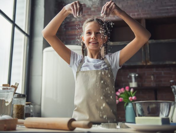 Cute little girl in apron is playing with flour and smiling while cooking in kitchen at home