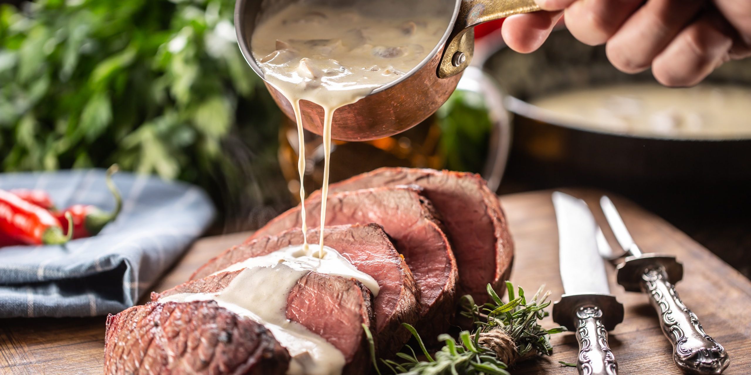 Rump steak cut to slices on a chopping board next to a knife and rosemary, with a mushroom sauce poured over it