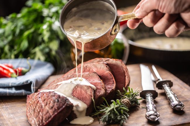 Rump steak cut to slices on a chopping board next to a knife and rosemary, with a mushroom sauce poured over it