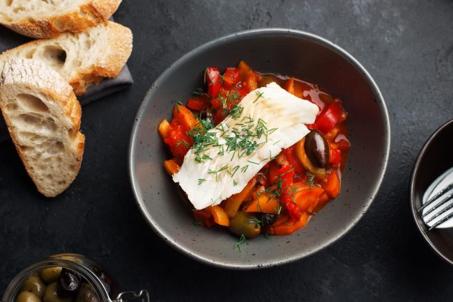 Delicious fish stew in paprika, tomatoes and white wine. Cod fillet white sea fish with organic vegetables, olives, onions, carrots, tomatoes, capsicum in a baking dish for lunch. Top view.