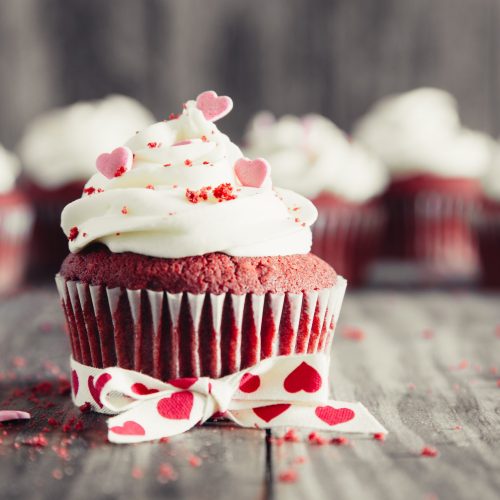 Red velvet cupcakes decorated with cheese frosting, heart shaped candies and crumbs on a rustic wooden table. Valentines dessert