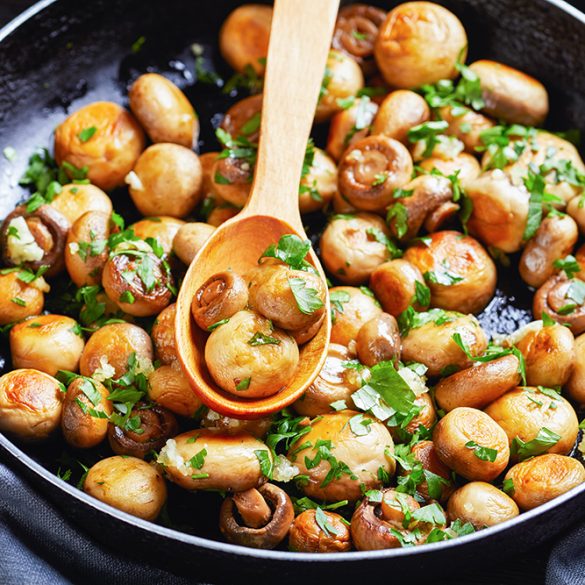 Sauteed in olive oil garlic small champignons with chopped parsley and lemon juice called al ajillo in Spain served on a frying pan on a dark wooden background with a spoon, top view, close-up