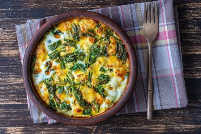 Ceramic bowl with vegetable frittata, simple vegetarian food. Frittata with egg, pepper, onion, cheese and green wild garlic leaves on table