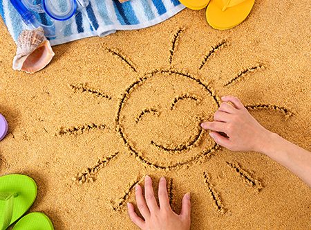 Child drawing a smiley sun in sand with towel, seashells and flip flops