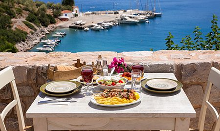 A table served for two with chicken souvlaki french fries, greek salad, snacks and drinks on the summer terrace of the hotel room by the seascape, beautiful summer greek holidays concept. Daylight.