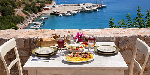 A table served for two with chicken souvlaki and french fries, greek salad, snacks and drinks on the summer terrace of the hotel room by the seascape, beautiful summer greek holidays concept.