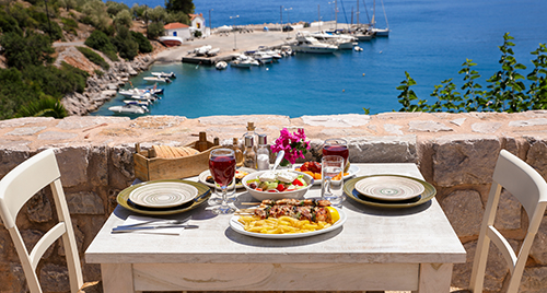 A table served for two with chicken souvlaki and french fries, greek salad, snacks and drinks on the summer terrace of the hotel room by the seascape, beautiful summer greek holidays concept.