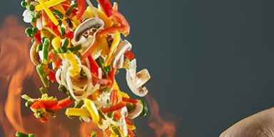 Frying pan wok with vegetables in the air on fire. Vegetables wok – peppers, onions, peas, mushrooms, beans, broccoli, zucchini. Asian food. Chinese food. The effect of levitation